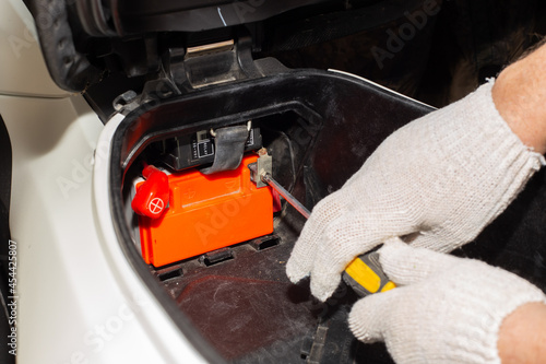 A gloved auto mechanic with a screwdriver unscrews the motorcycle battery terminals for repair or replacement. Car service in a car service.
