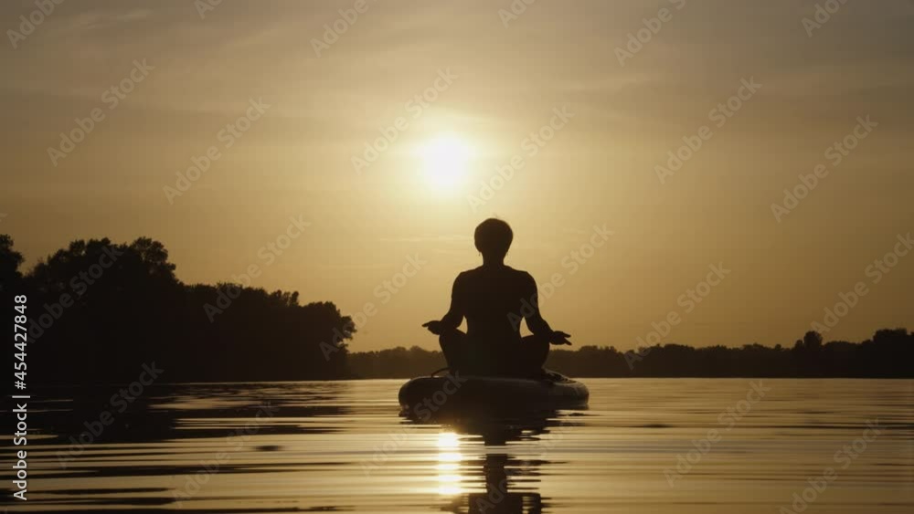 Silhouette of woman sitting in lotus pose on SUP board and meditating on water, sun setting on background. Relaxing and enjoying harmony of nature. Healthy lifestyle. Concept of recreation Silhouette of woman sitting in lotus pose on SUP board and meditating on water, sun setting on background. Relaxing and enjoying harmony of nature. Healthy lifestyle. Concept of recreation