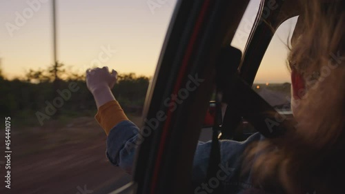 woman in car holding hand out window feeling wind blowing through fingers driving in countryside on road trip travelling for summer vacation enjoying freedom on the road at sunrise photo