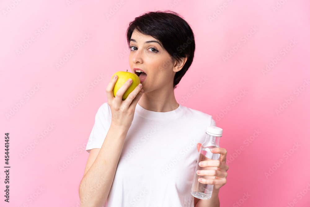 Woman with short hair isolated on pink background with a bottle of water and eating an apple Woman with short hair isolated on pink background with a bottle of water and eating an apple