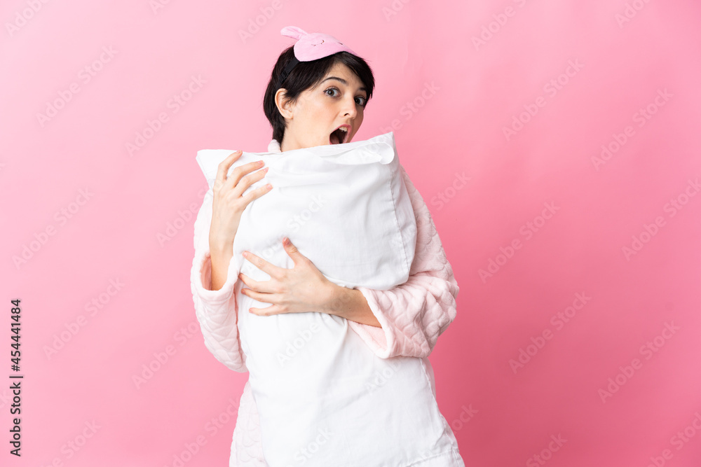 Woman with short hair isolated on pink background in pajamas and holding a pillow with happy expression Woman with short hair isolated on pink background in pajamas and holding a pillow with happy expression