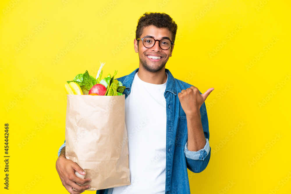 Young Brazilian man holding a grocery shopping bag isolated on yellow background pointing to the side to present a product Young Brazilian man holding a grocery shopping bag isolated on yellow background pointing to the side to present a product
