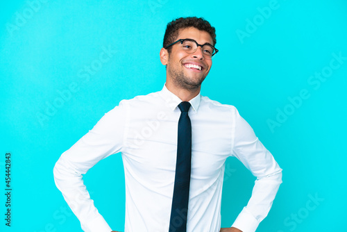 Young business Brazilian man isolated on blue background posing with arms at hip and smiling
