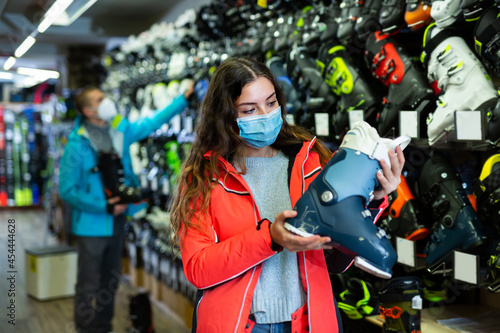 Young customer woman in protective mask is choosing boots for skiing in store