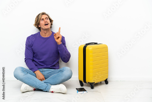 Young blonde man with suitcase sitting on the floor thinking an idea pointing the finger up