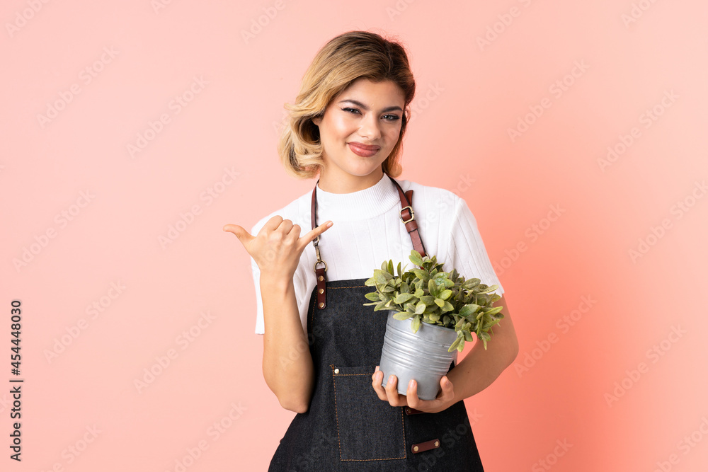 Russian gardener girl holding a plant isolated on pink background making phone gesture Russian gardener girl holding a plant isolated on pink background making phone gesture