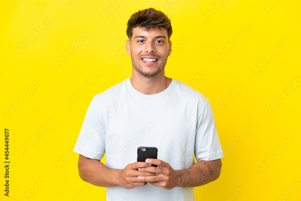 Young caucasian handsome man isolated on yellow background looking at the camera and smiling while using the mobile Young caucasian handsome man isolated on yellow background looking at the camera and smiling while using the mobile