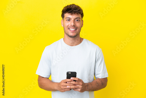 Young caucasian handsome man isolated on yellow background looking at the camera and smiling while using the mobile © luismolinero