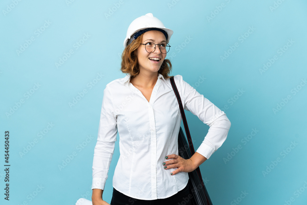 Young architect Georgian woman with helmet and holding blueprints over isolated background posing with arms at hip and smiling