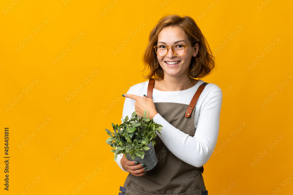 Young Georgian woman holding a plant isolated on yellow background pointing to the side to present a product Young Georgian woman holding a plant isolated on yellow background pointing to the side to present a product