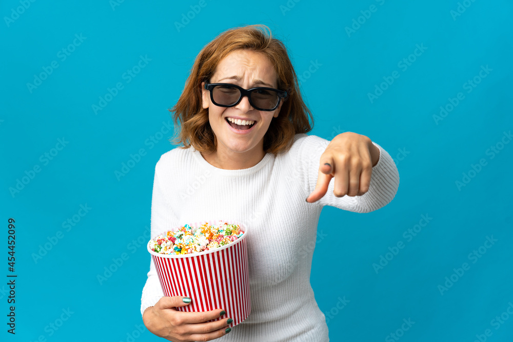 Young Georgian woman isolated on blue background with 3d glasses and holding a big bucket of popcorns while pointing front