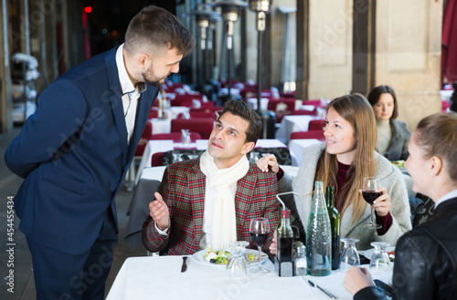 Positive elegant company of three sitting on terrace restaurant, talking with manager.