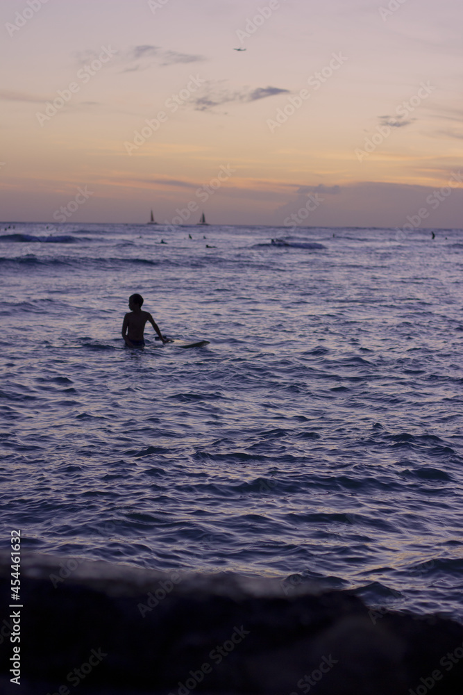 Silhouette of surfer at dusk with calm water and sailboats in background
