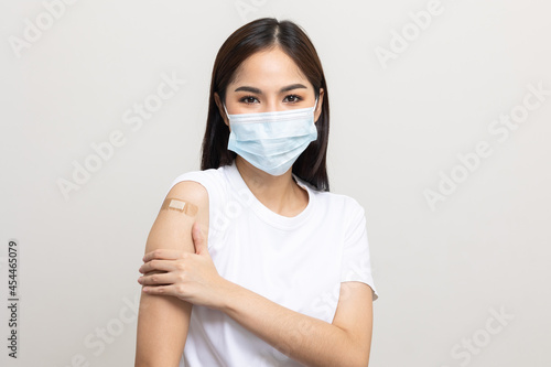 Young beautiful asian woman wearing mask and getting a vaccine protection the coronavirus. Happy female showing arm with bandage after receiving vaccination on isolated white background.