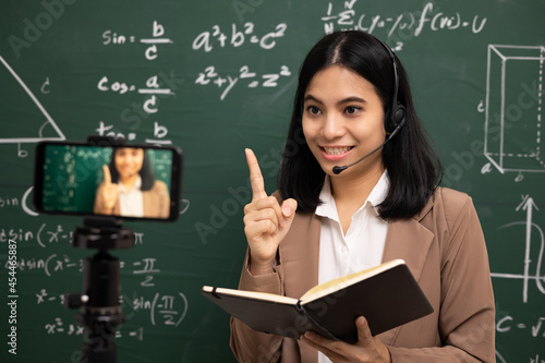 Young asian teacher woman standing looking at camera and video conference with student. Female teacher training the mathematics in classroom from live stream with smartphone online course.