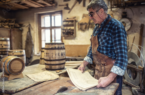 Aged craftsman reads drawings in his rustic wooden barrels producing workshop
