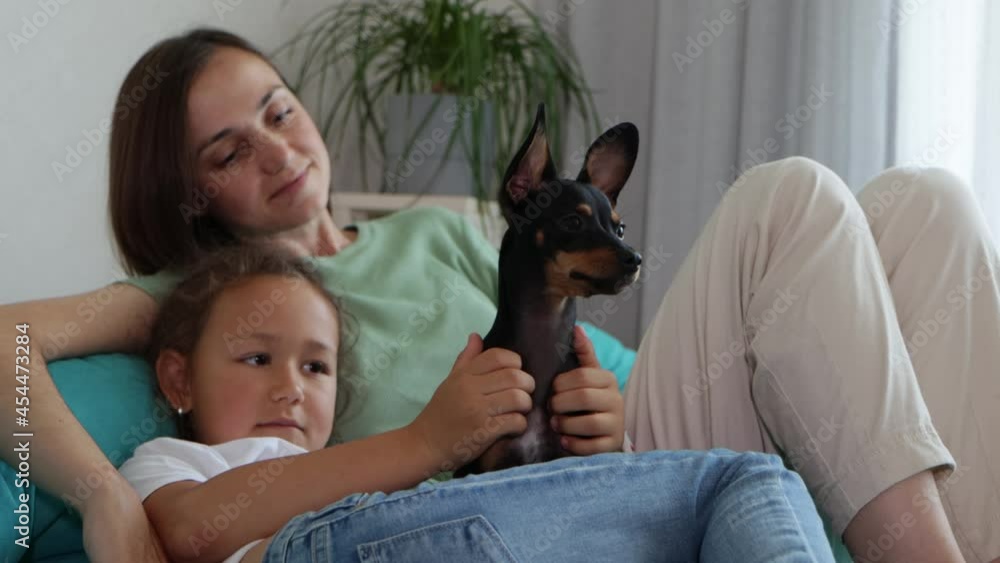 Mother and daughter having a good time with their dog at sofa. Portrait of black toy terrier with owners at home Mother and daughter having a good time with their dog at sofa. Portrait of black toy terrier with owners at home