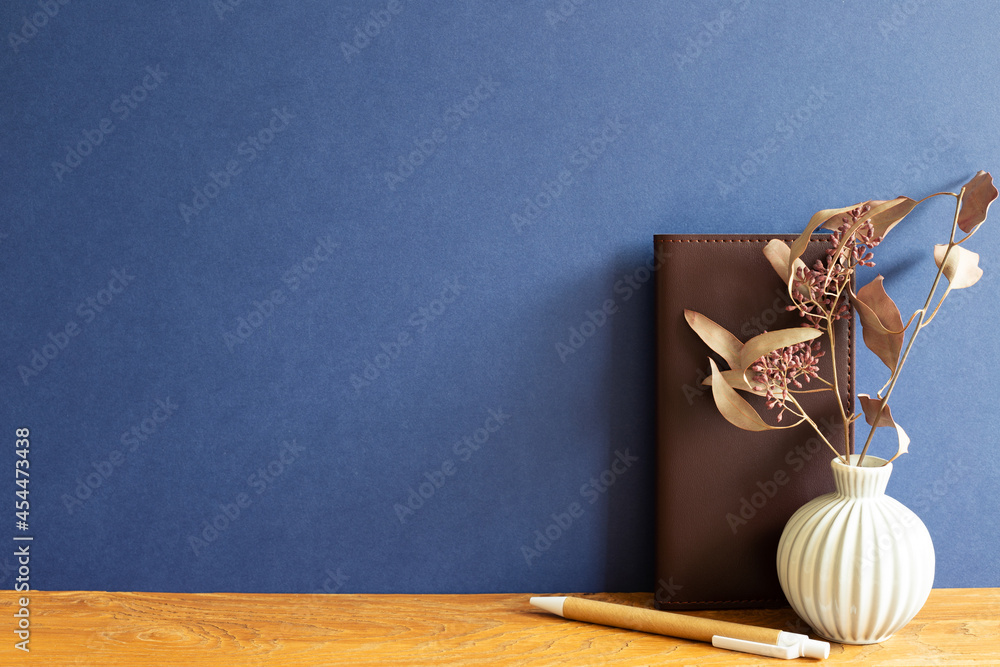 Brown leather notebook, pen, dry flowers on wooden desk. blue wall background