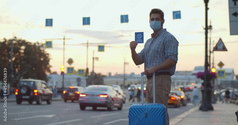 Man traveler in safety mask with suitcase standing on street and holding vaccine passport Man traveler in safety mask with suitcase standing on street and holding vaccine passport