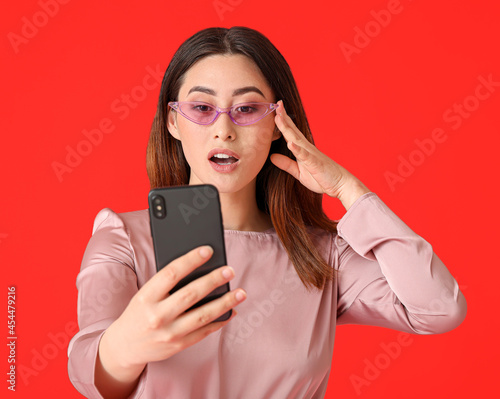 Stylish young Asian woman with mobile phone on red background