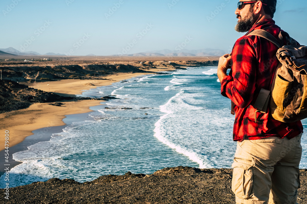 Rear view of man standing with backpack and enjoying the wonderful landscape in front of him with beach and coast - adult people and alternative walking vacation free and alone