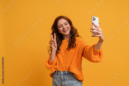 Attractive red head woman in stylish fall outfitm sweater and jeans posing over yellow background in studio. Making self portrait by smartphone.