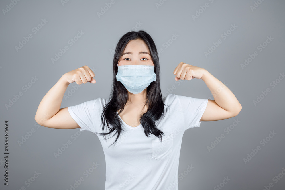 Portrait of healthy strong Caucasian Asian woman in medical face mask to protect Covid-19 (Coronavirus) triumphing with raised fists on gray background Portrait of healthy strong Caucasian Asian woman in medical face mask to protect Covid-19 (Coronavirus) triumphing with raised fists on gray background
