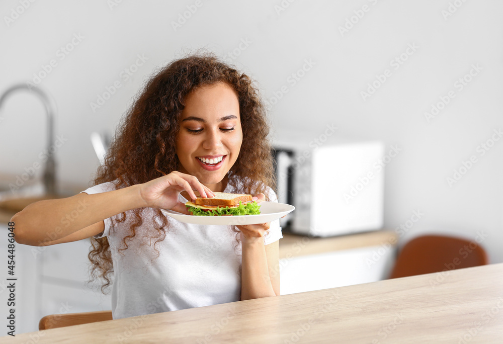 Young African-American woman eating tasty sandwich in kitchen Young African-American woman eating tasty sandwich in kitchen