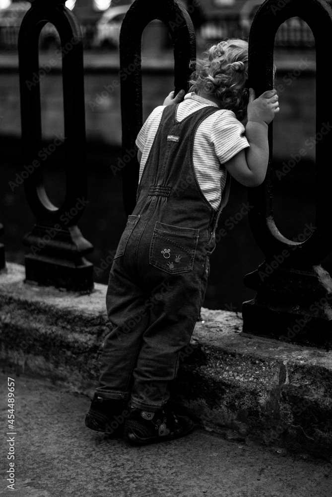 Fototapeta premium Little curly-haired boy in denim overalls looks over the fence at the river
