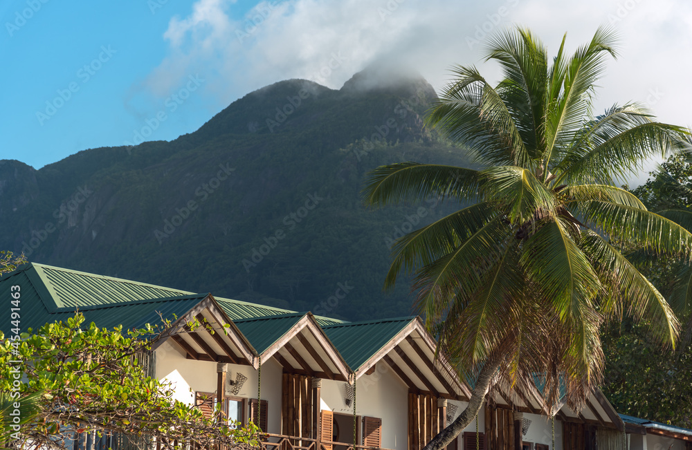 Scenic view at tropical landscape with high palm tree, tropical villa and mountain against blue sky. Seychelles, Mahe island, Beau Vallon beach