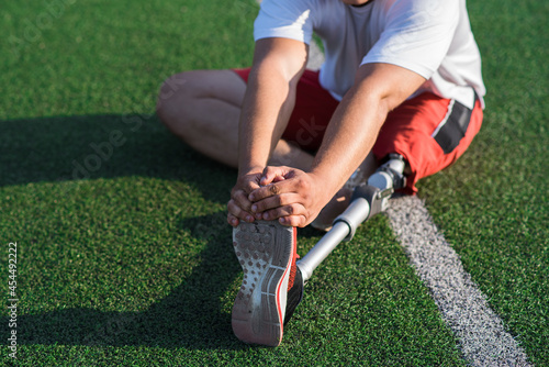Close up of a Caucasian guy with a prosthetic leg stretching while sitting on the field at the stadium. Sport concept.