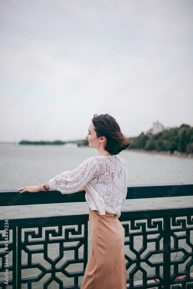 A happy girl on the river bank, standing on the bridge, smiling, a girl walks on the bridge by the river. Freedom. The wind is blowing the girl's hair. A happy girl on the river bank, standing on the bridge, smiling, a girl walks on the bridge by the river. Freedom. The wind is blowing the girl's hair.