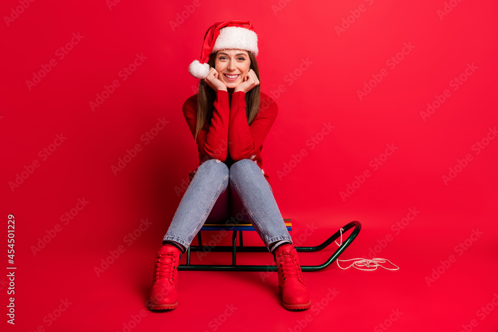Full size photo of nice young lady sit wear holiday sweater cap jeans boots isolated on red color background Full size photo of nice young lady sit wear holiday sweater cap jeans boots isolated on red color background