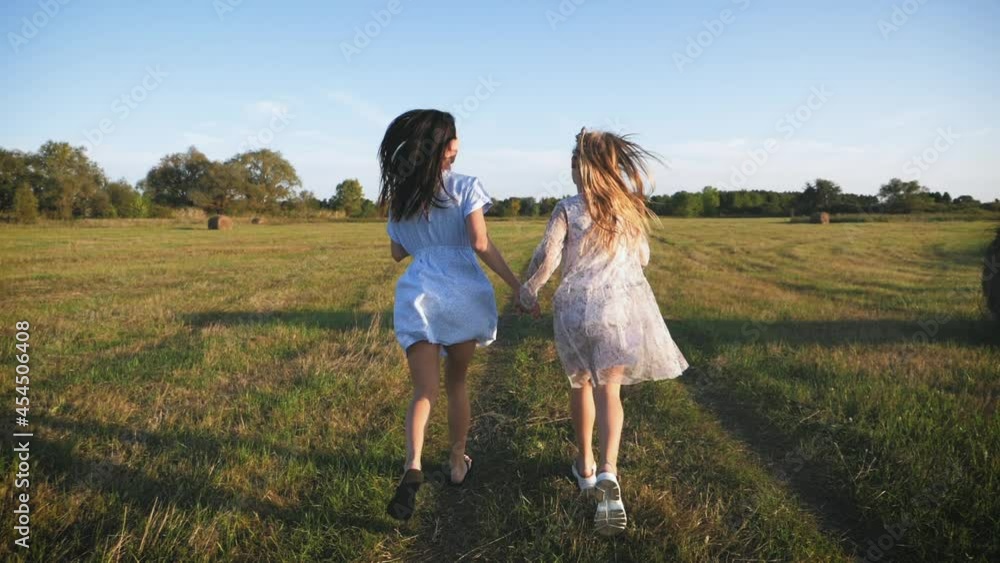 Friendly family. Mother and daughter holding hands at sunset running in the meadow. Happy mom and daughter walk holding hands towards the sunset in summer. Freedom happiness concept. Friendly family. Mother and daughter holding hands at sunset running in the meadow. Happy mom and daughter walk holding hands towards the sunset in summer. Freedom happiness concept.