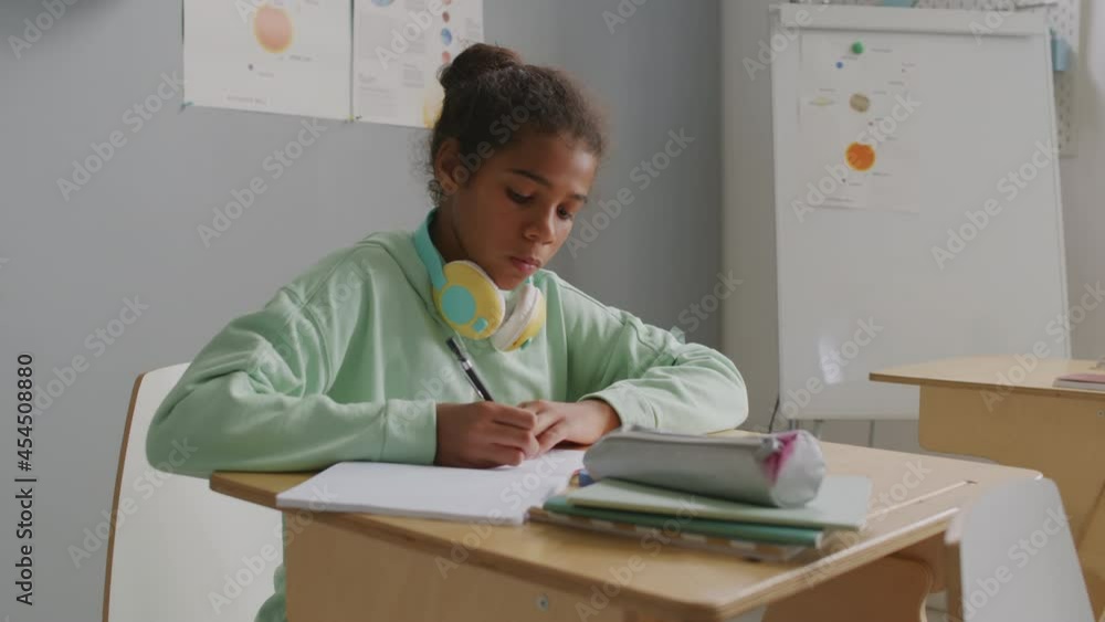 Handheld shot of African-American 11-year-old girl sitting at desk in classroom and writing in her notebook during lesson Handheld shot of African-American 11-year-old girl sitting at desk in classroom and writing in her notebook during lesson
