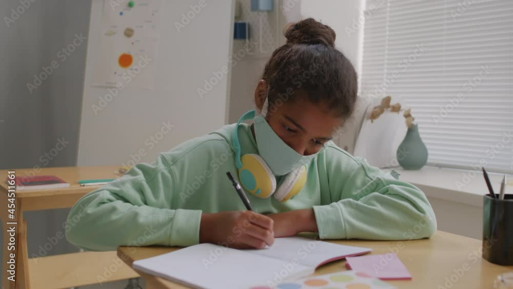 Handheld shot of African-American 11-year-old girl in face mask sitting at desk in classroom and writing in notebook during lesson Handheld shot of African-American 11-year-old girl in face mask sitting at desk in classroom and writing in notebook during lesson