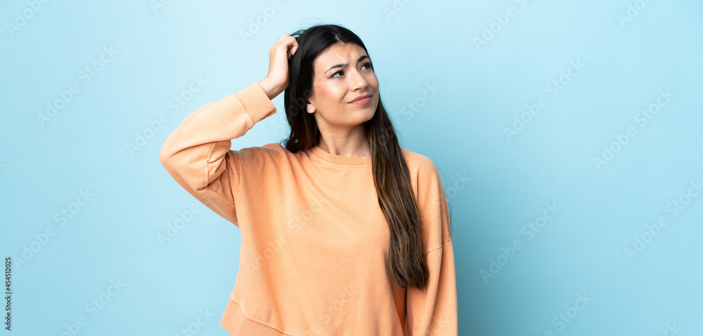 Fototapeta premium Young brunette girl over isolated blue background having doubts while scratching head