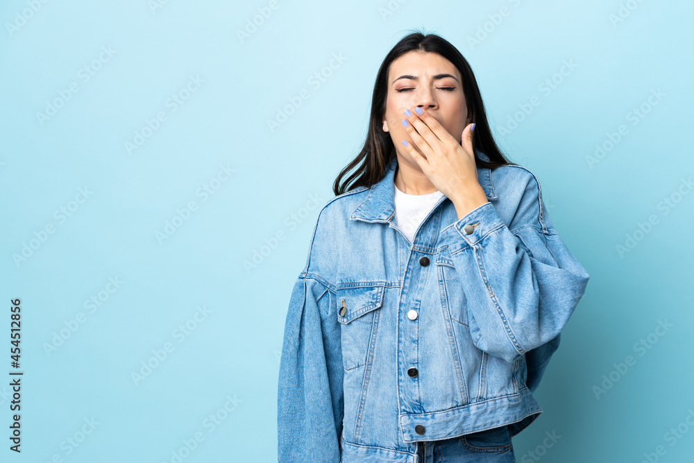 Young brunette girl over isolated blue background yawning and covering wide open mouth with hand Young brunette girl over isolated blue background yawning and covering wide open mouth with hand