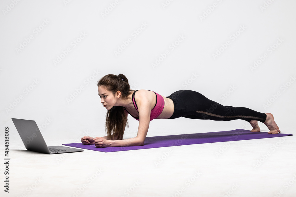 Strong sporty woman is doing working out on yoga mat in front of her laptop, wearing sport outfit on white background.