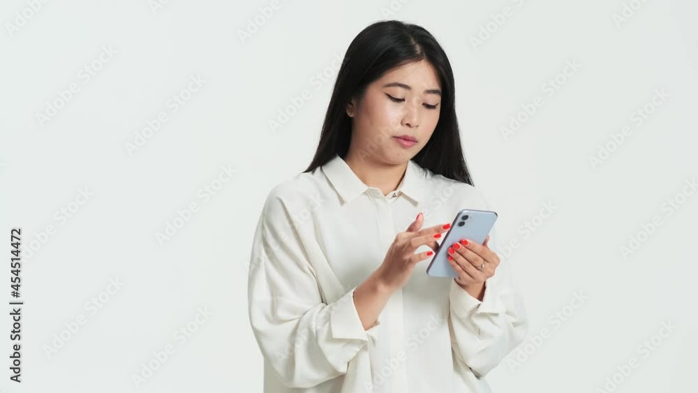Positive asian korean woman using her phone standing isolated over white background in the studio Positive asian korean woman using her phone standing isolated over white background in the studio