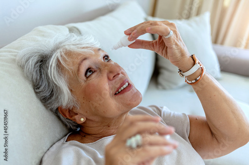 A sick old woman suffering from irritated eye, optical symptoms. A senior woman applying eye drop by herself. A woman dropping eye drop medicine healing her eye pain. Health care and medicine concept. photo