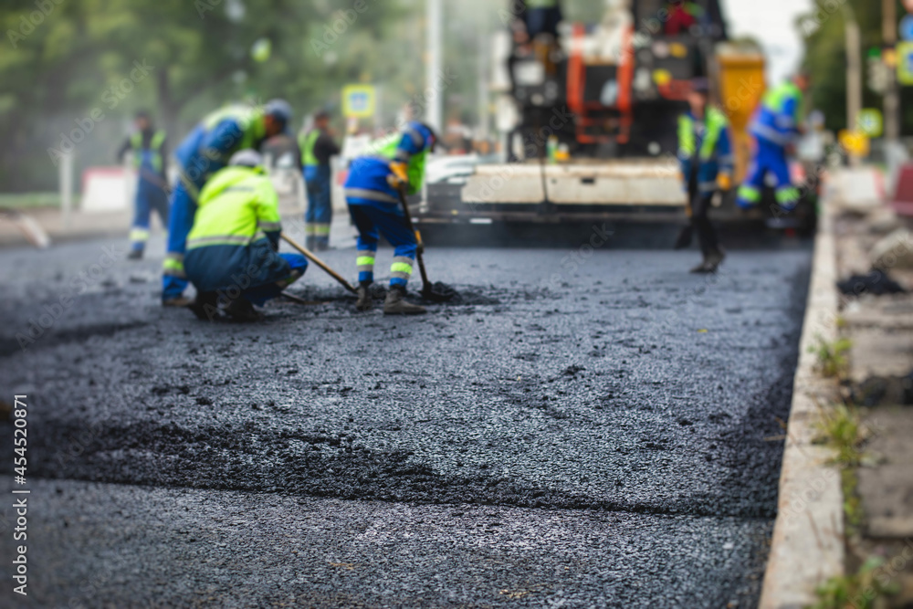 Process of asphalting, blacktopping and paving, group of workers with shovels finishing the asphalt layer, with asphalt paver machine and steam roller compactor vehicle in the background