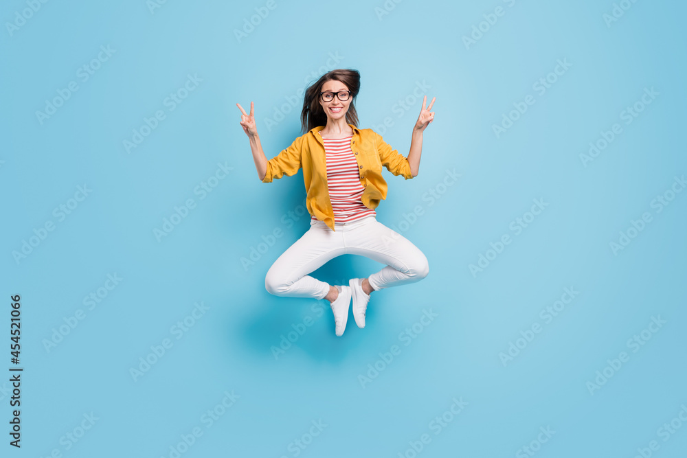 Full body photo of young woman happy positive smile show peace cool v-sign jump up isolated over blue color background Full body photo of young woman happy positive smile show peace cool v-sign jump up isolated over blue color background