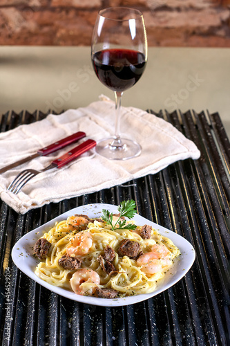 Spaghetti pasta with white sauce, meat, shrimp, mozzarella cheese and fresh basil on a white plate, red wine, on a black grill and rustic wall