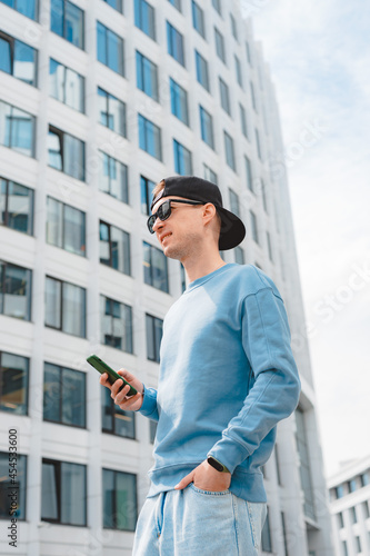 Positive hipster guy with backpack standing outdoor holding smartphone while looking away with smile