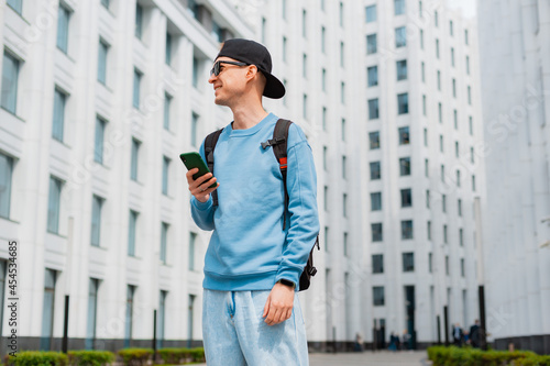 Positive hipster guy with backpack standing outdoor holding smartphone while looking away with smile © shangarey