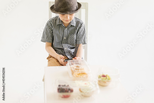 A young boy in a shirt and hat sits in front of cakes with a phone in his hands.