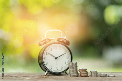 Alarm clock with step of coins on wood table with green nature background 