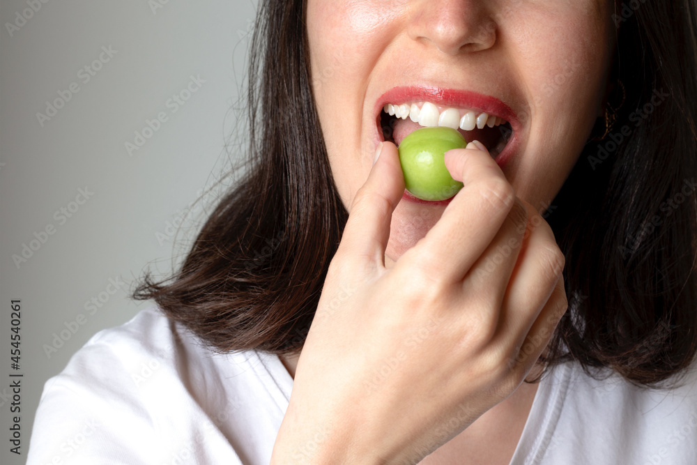 a young woman eating a green sour plum with blurred grey background. Selective Focus. The young woman's hand is holding a sour green plum to eat. Healthy nutrition a young woman eating a green sour plum with blurred grey background. Selective Focus. The young woman's hand is holding a sour green plum to eat. Healthy nutrition