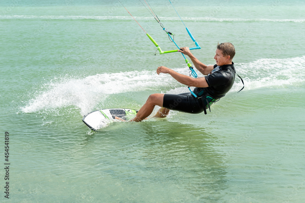Fototapeta premium Young adult caucasian fit male person enjoy riding kite surf board in sun uv protection suit on bright sunny day against blue sky at sea or ocean shore. Watersport adrenaline fun adventure acitivity